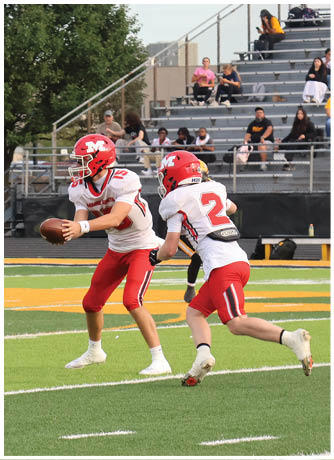 Three people in red and white football uniforms are on a field with one person holding a football preparing to pass while the others are positioned nearby. Description generated by AI