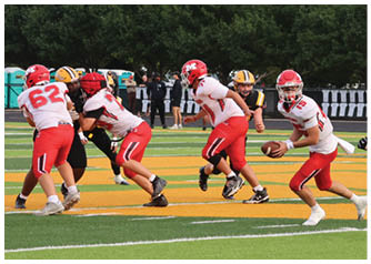 Players in red and white uniforms are engaged in a football play on a green and yellow field with trees and spectators in the background. Description generated by AI