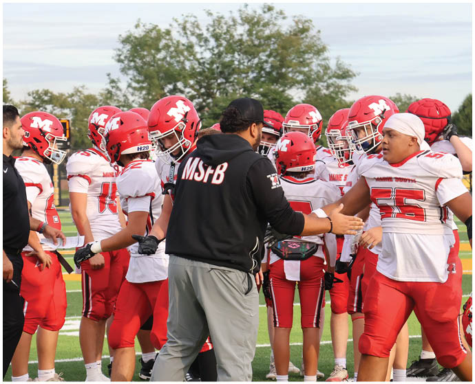 A group of people in red and white football uniforms are gathered on a field with one person in a black jacket appearing to give instructions. Description generated by AI