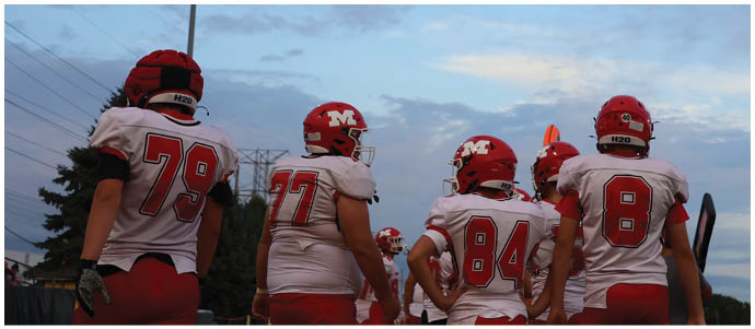 Four people wearing red helmets and white football uniforms with numbers on the back are standing outdoors under a partly cloudy sky. Description generated by AI