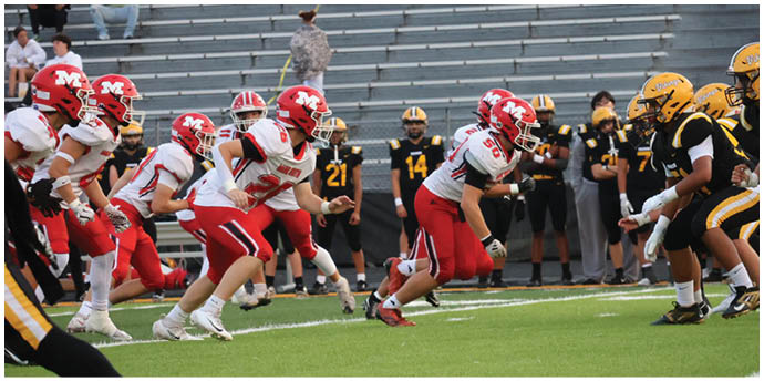 Two opposing American football teams in red and white and black and yellow uniforms are lined up facing each other on a field with empty bleachers in the background. Description generated by AI