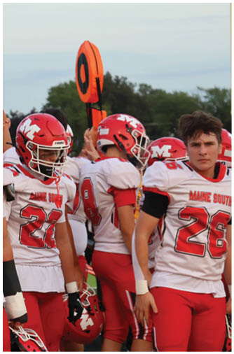 People wearing red and white football uniforms and helmets are standing together on a field during a game or practice. Description generated by AI
