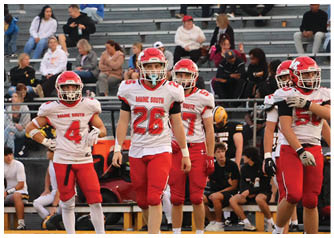 Several people in red and white football uniforms stand on a field with a crowd of spectators seated on bleachers in the background. Description generated by AI
