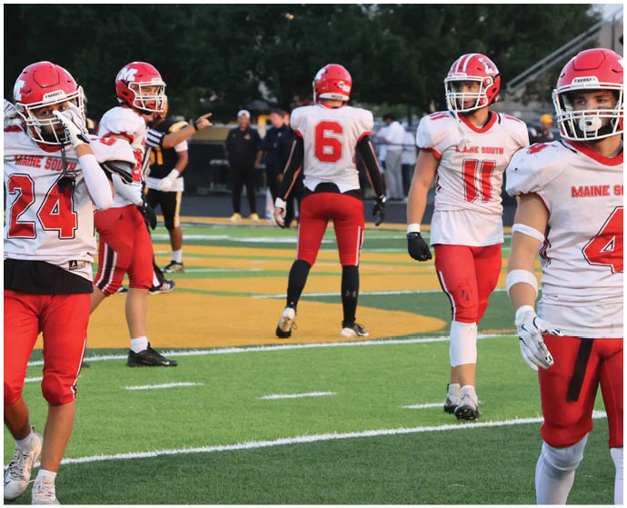 People wearing red and white football uniforms are standing on a green and yellow field, some facing forward and others with their backs turned. Description generated by AI