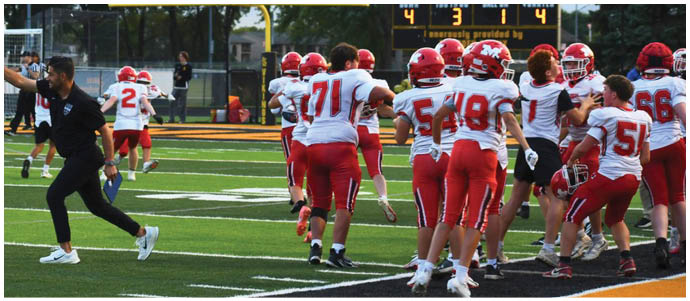 Adults in football uniforms are on a field near a scoreboard showing a game in progress with a referee signaling and players in red and white uniforms gathered. Description generated by AI