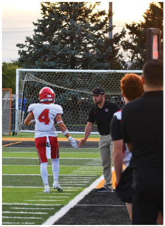 People in football uniforms and a referee stand on a field near the end zone with a scoreboard showing zero. Description generated by AI