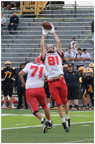 Two athletes in red and white uniforms jump to catch a football while other players and spectators watch from the sidelines and bleachers of a sports field. Description generated by AI