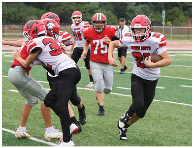 People wearing football uniforms are actively playing on a field with one person running while holding the ball and others chasing or blocking. Description generated by AI
