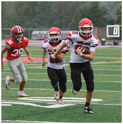 Two people wearing red helmets and white football jerseys are playing football on a rainy field with one person running while holding the ball and the other following behind. Description generated by AI
