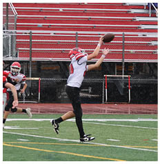 Children wearing helmets and football uniforms are playing on a wet outdoor football field near the sidelines. Description generated by AI