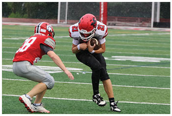 Three people wearing red helmets and sports uniforms are playing football on a green field with white yard lines and goalposts in the background. Description generated by AI