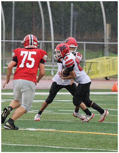 Two people in football uniforms are engaged in a play on a rainy field while a third person in a red jersey approaches. Description generated by AI