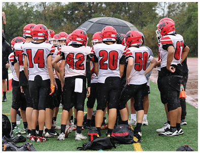 A group of people wearing red helmets and white sports jerseys with large red numbers are standing together on a sports field. Description generated by AI
