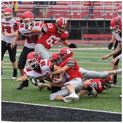 Several people wearing red and white football uniforms are engaged in a tackle near the end zone on a rainy field. Description generated by AI