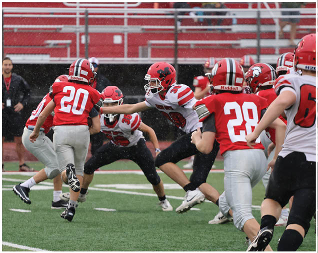 Several people wearing football uniforms are actively engaged in a play on a rainy field with empty bleachers in the background. Description generated by AI