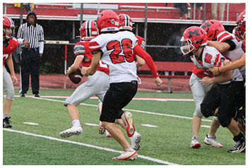 Several people wearing red and white football uniforms are actively playing on a green field with a red and black bleacher background. Description generated by AI