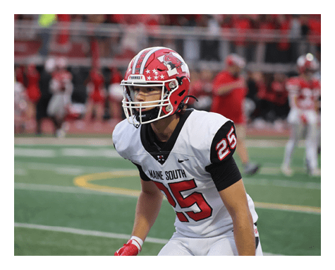 A person wearing a red and white football helmet and a white uniform with the number 25 is standing on a football field in a ready position with a blurred crowd in the background. Description generated by AI