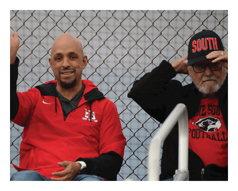 A person wearing a red jacket is smiling and waving while sitting next to another person in a black jacket and a black cap with red lettering, both seated in front of a chain-link fence. Description generated by AI