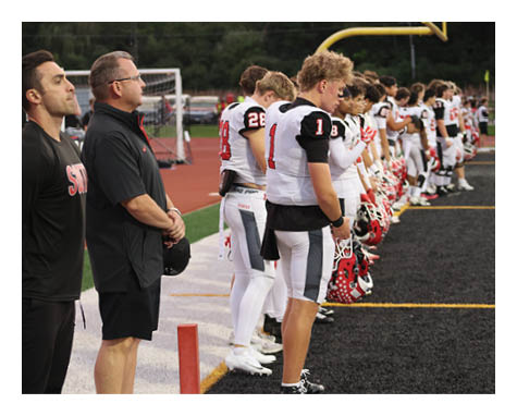 People in football uniforms and coaches stand in a line on the sidelines of a football field facing the field, appearing attentive and respectful. Description generated by AI