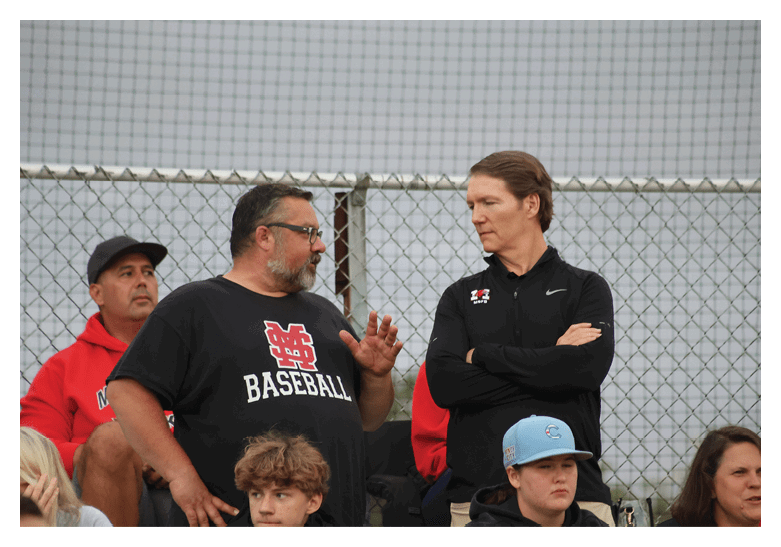 Three adults stand near a chain-link fence with one person in a black baseball shirt gesturing while talking to another person in a black jacket with crossed arms. Description generated by AI