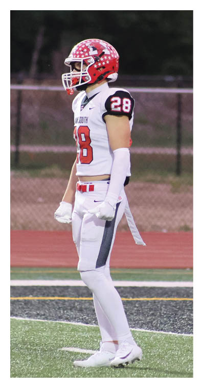 A person wearing a red helmet and white football uniform with the number 28 stands on a football field near the sideline. Description generated by AI