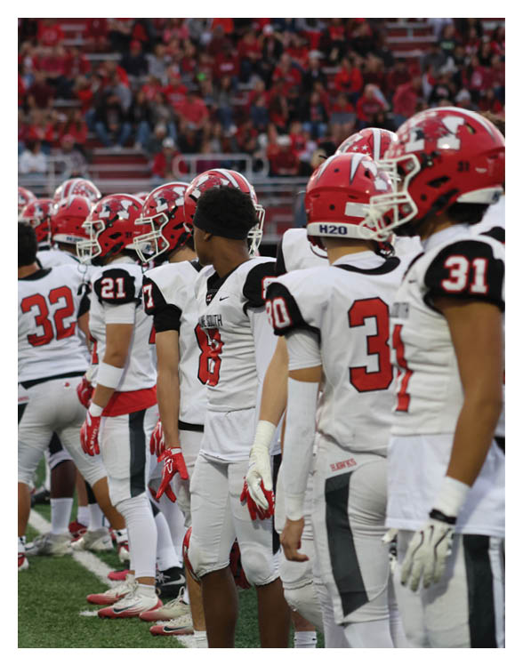 A group of people wearing white and red football uniforms stand in a line on a field, some looking forward and others looking to the side. Description generated by AI