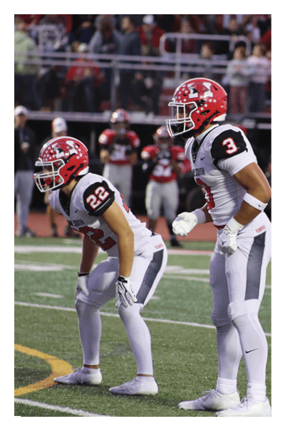 A person wearing a red helmet and white football uniform with the number 3 is standing on a football field in a ready position. Description generated by AI