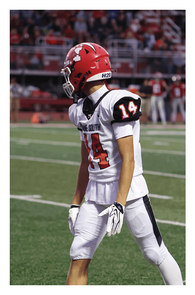 A person in white athletic gear is on a football field with a referee and other people in the background near the stands. Description generated by AI