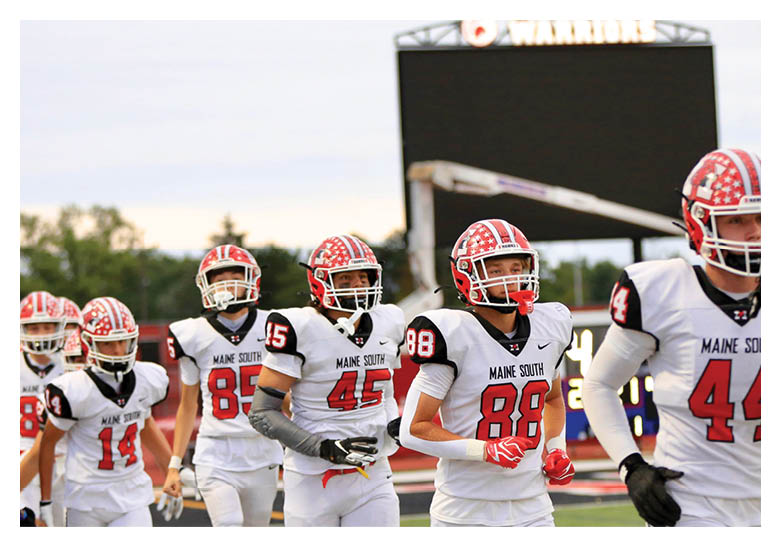 A group of people wearing white and red football uniforms with helmets are walking together on a field with a scoreboard in the background. Description generated by AI