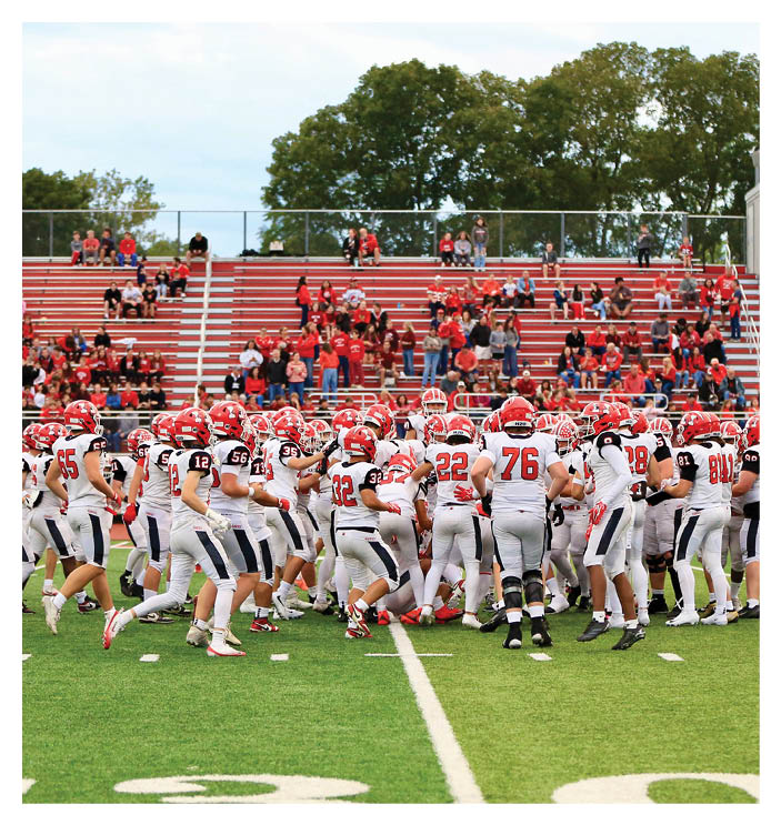A group of football players in white uniforms with red helmets are gathered on a green field near the 20-yard line with spectators in red clothing seated in the bleachers behind them. Description generated by AI