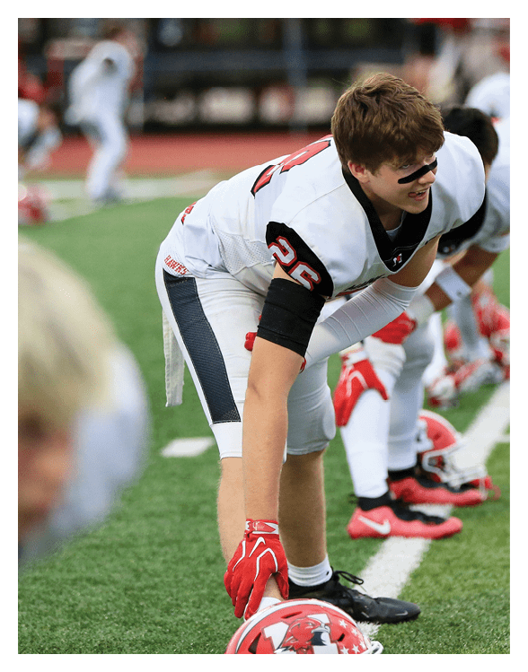 A person in a white and black football uniform with red gloves is crouched on a green field, preparing for a play. Description generated by AI