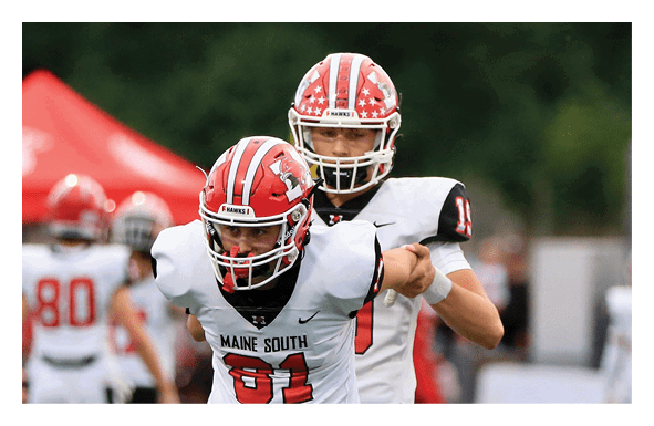 Five people wearing matching red and black cheerleading uniforms with “Maine South" on the front are standing close together outdoors with trees and a sports field in the background. Description generated by AI