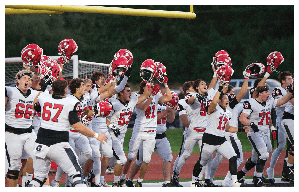 A group of people wearing white sports uniforms enthusiastically raise red helmets in celebration on a football field near a goalpost. Description generated by AI