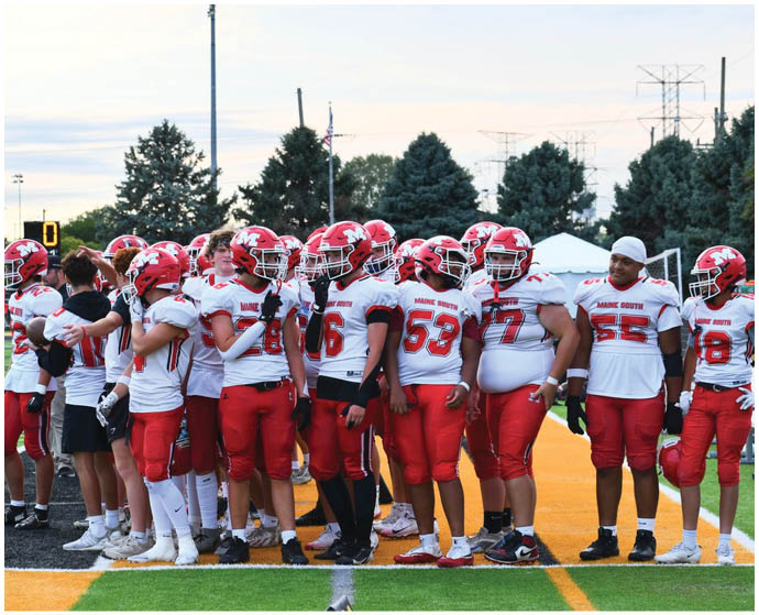 A group of people wearing red and white football uniforms stand together on the sidelines of a sports field with trees and power lines in the background. Description generated by AI