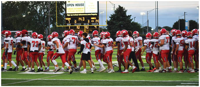 A group of people wearing red and white football uniforms are gathered on a field near a scoreboard displaying game information and an announcement for an open house event. Description generated by AI
