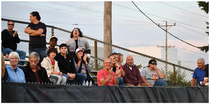 A group of people sitting and standing on bleachers outdoors, some clapping and others watching attentively. Description generated by AI
