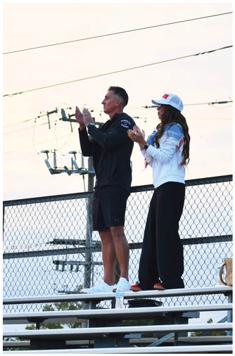 Two adults stand on metal bleachers outdoors at sunset, clapping and looking toward the left, with one wearing a white jacket and cap and the other in a black outfit. Description generated by AI
