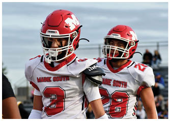 Two people wearing red football helmets and uniforms stand outdoors with a blurred background. Description generated by AI