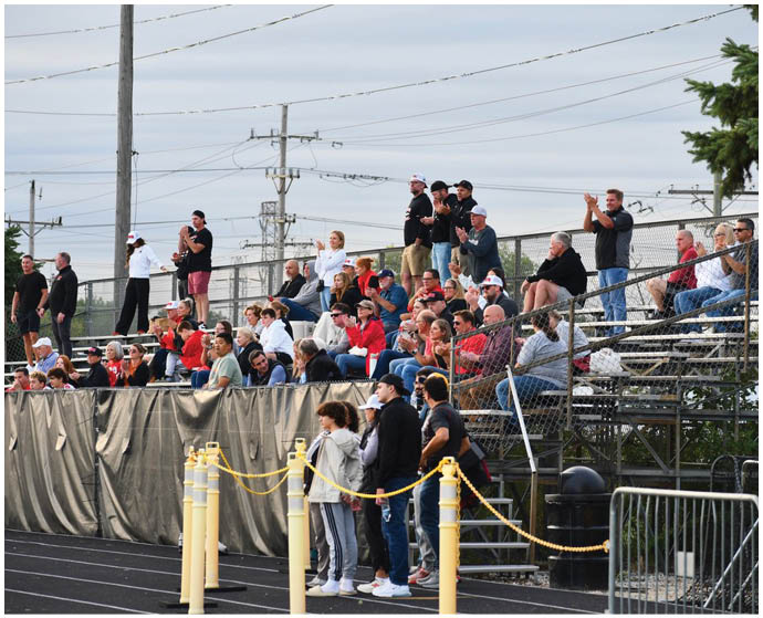 A crowd of people wearing casual clothing is gathered on bleachers and standing near a track, watching an event with some clapping and others focused on the activity. Description generated by AI