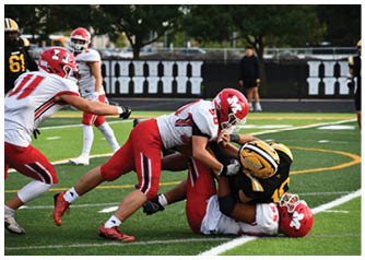 Three football players in red and white uniforms tackle a player in a black and yellow uniform on a green field during a game. Description generated by AI