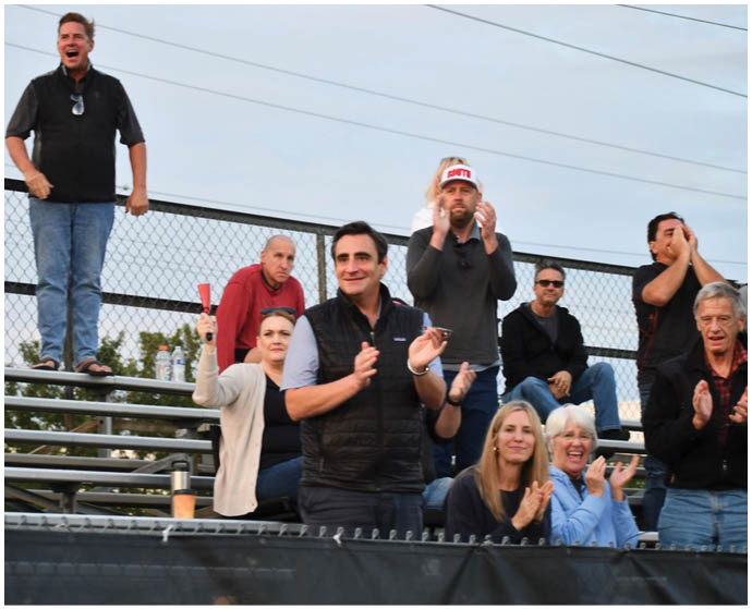 People in casual clothing are standing and sitting on bleachers outdoors, clapping and cheering. Description generated by AI