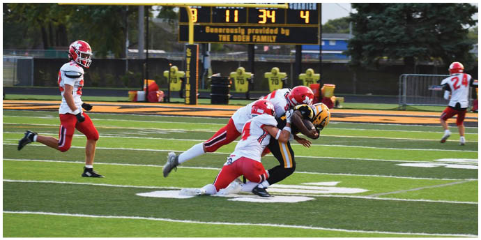 A group of people in football uniforms are playing on a field with one person in a black and yellow uniform being tackled by two people in white and red uniforms near the 10-yard line with a scoreboard in the background. Description generated by AI