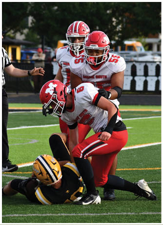 Two people in football uniforms are on a field with one person in black and yellow lying on the ground and the other in red and white leaning over them while other players and a referee stand nearby. Description generated by AI