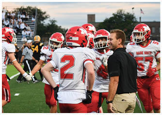 A group of football players in red and white uniforms are gathered on a field with a coach in a black shirt and khaki pants during a game. Description generated by AI