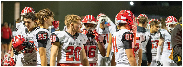 A group of people wearing white and red football uniforms are standing together on a field, some holding helmets and others engaged in conversation. Description generated by AI