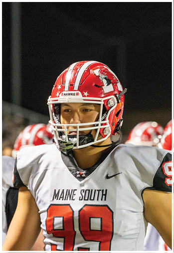 A person wearing a red football helmet with a hawk logo and a white jersey that says “Maine South" stands on a field at night. Description generated by AI