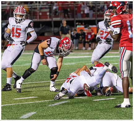 Players in red and white football uniforms are engaged in a tackle on a green field with spectators in the background. Description generated by AI