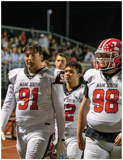 Three people in white football uniforms with red numbers are walking on a field with a crowd in the background. Description generated by AI
