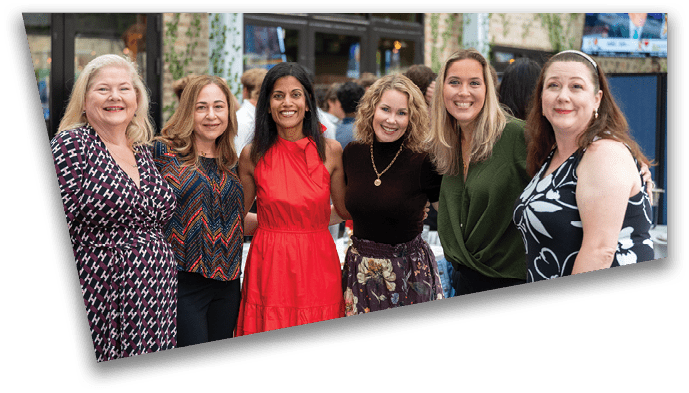 Six adults smiling and standing close together in a casual indoor setting with plants and windows in the background. Description generated by AI