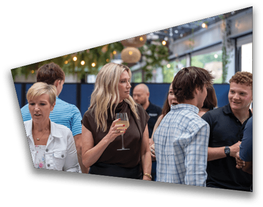 A group of people are socializing indoors with one person holding a glass of wine and others engaged in conversation under hanging lights and greenery. Description generated by AI